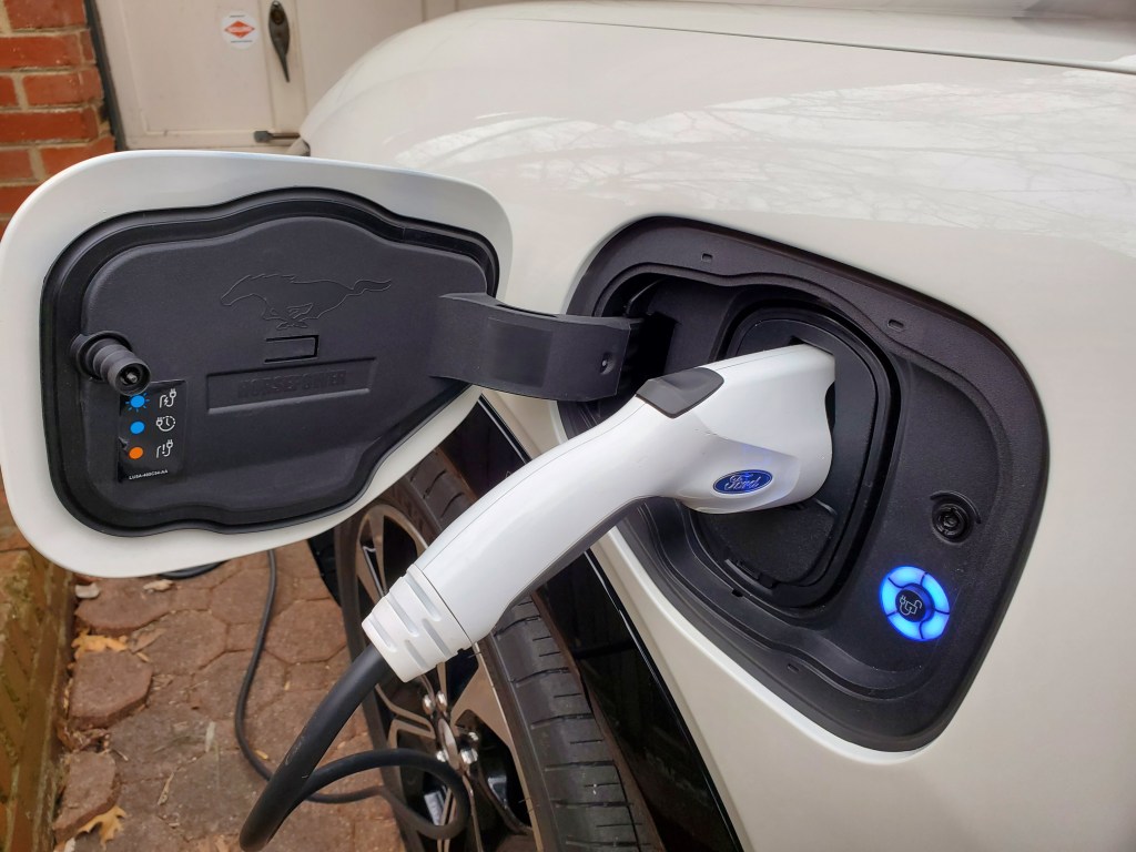 Ford MACH-E left-front fender-mounted charge port with charger plugged in and lit a Mustang logo on the inside of the charge port door